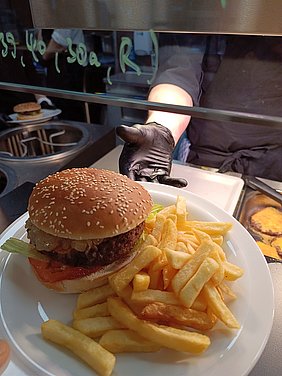 Foto Hand mit schwarzem Handschuh reicht Teller mit Burger und Pommes in Richtung Kamera