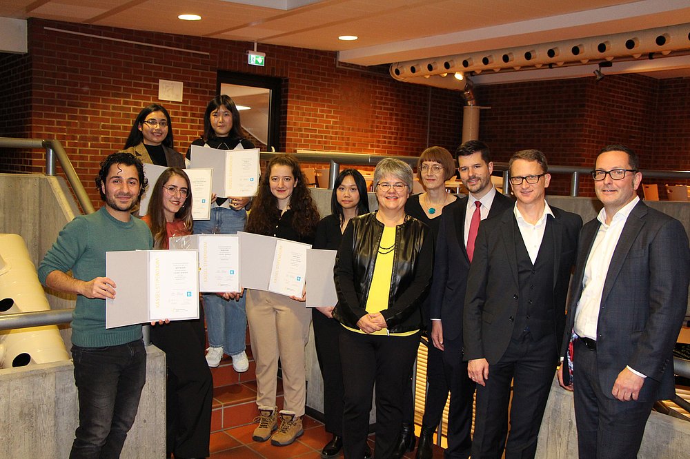 Photo: Eleven people standing in a semicircle in a building, six of them holding certificates in their hands. Photo: Eleven people standing in a semicircle in a building, six of them holding certificates in their hands.
