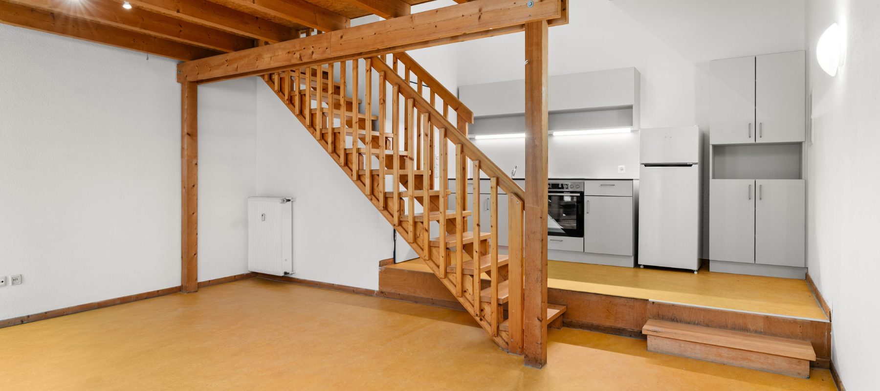 Foto vom Wohnbereich der Studio-Wohnung in Richtung Treppe und Küchenzeile. Zu sehen ist eine Holztreppe auf die zweite Ebene, dahinter ist eine Küchenzeile auf einem kleinen Podest mit Stufe.