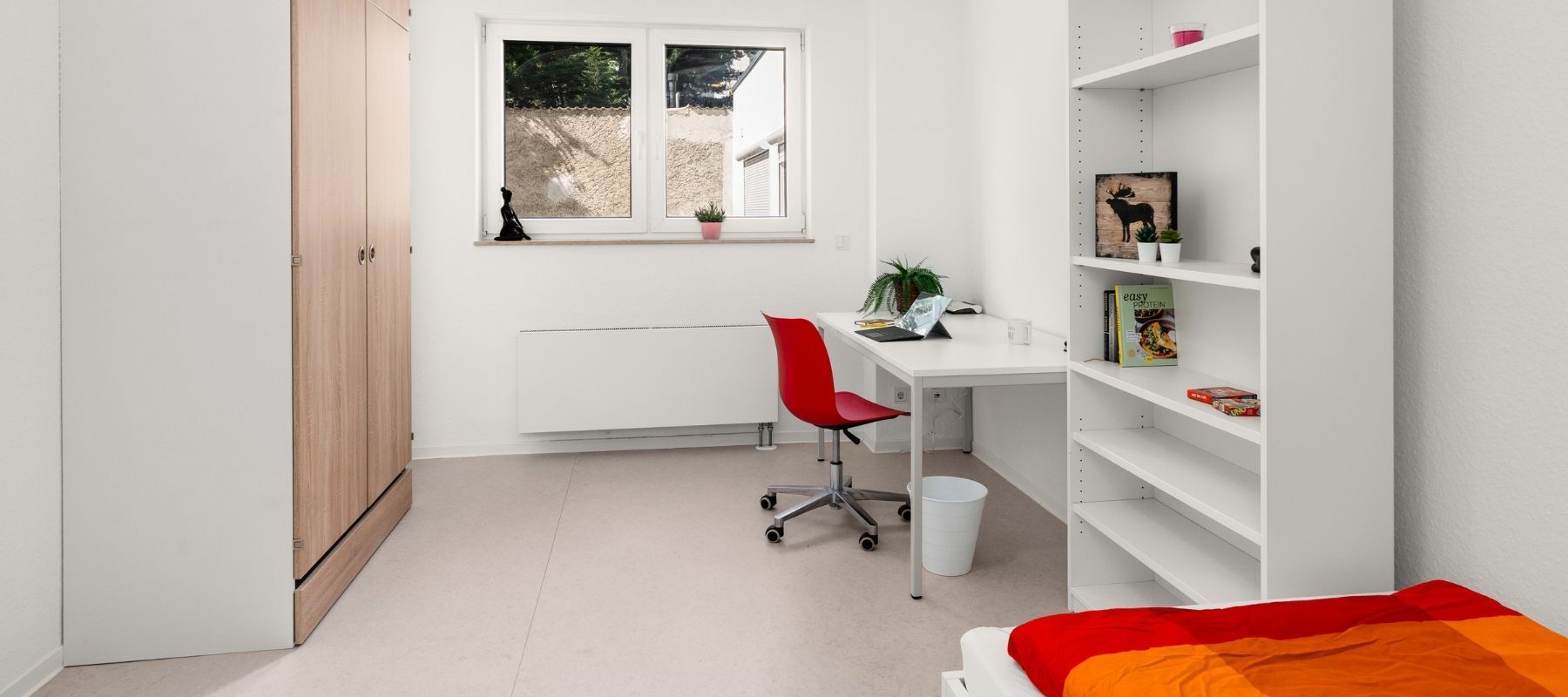 Foto eines Zimmeres in einem Doppelapartment. Zu sehen sind ein Kleiderschrank, ein Bücherregal sowie ein Schreibtisch mit Stuhl.
